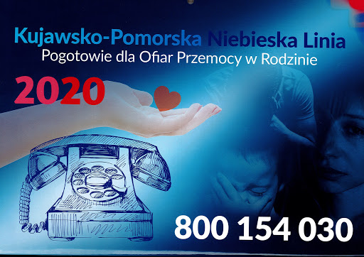 grafika przedstawiająca dłoń, trzymającą serce, pod którą znajduje się telefon oraz numer: 800 154 030, w tle widoczna schylona postać mężczyzny, trzymajacego twarz w dłoniach, głowa dziecka, które również zakrywa twarz dłonią, widać także smutną twarz kobiety, na górze grafiki napis: Kujawsko-Pomorska Niebieska Linia Pogotowie dla Ofiar Przemocy w Rodzinie