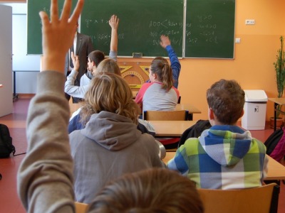 Zdjęcie przedstawiające dzieci podczas lekcji - widok od tyłu. Niektóre dzieci zgłaszają się do odpowiedzi, przed tablicą widoczny jest także nauczyciel.